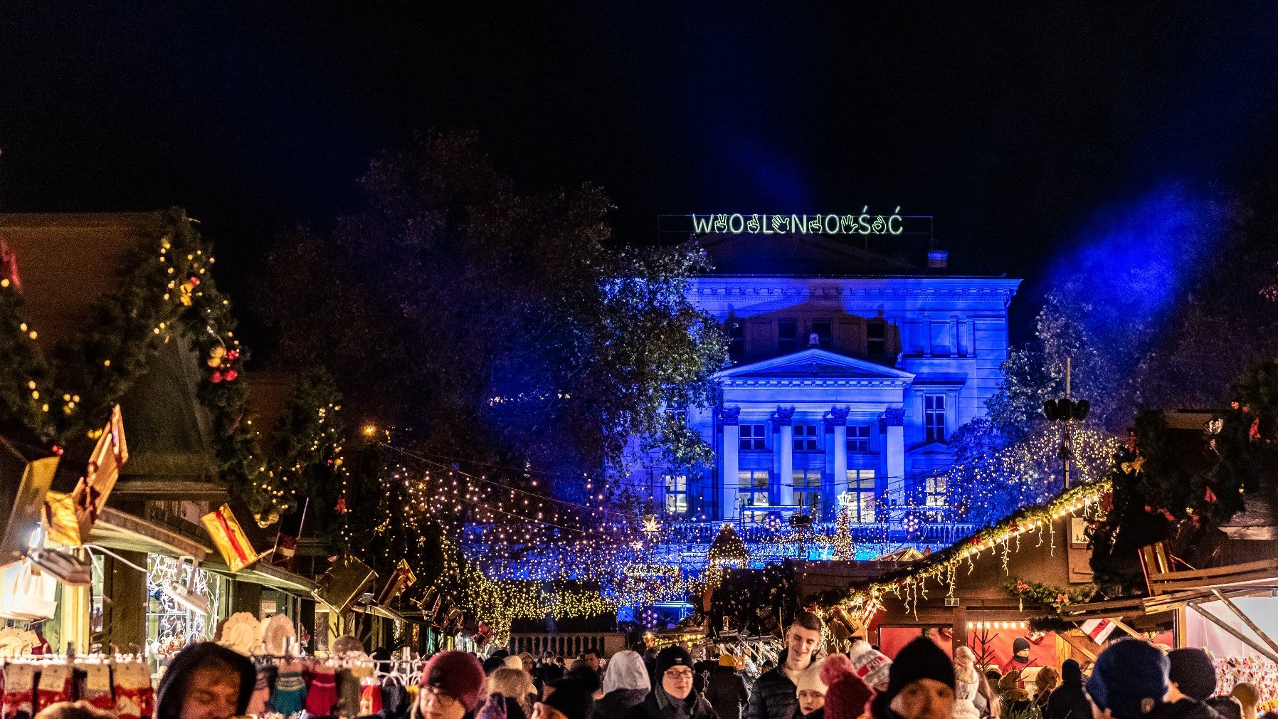 Jarmark na pl. Wolności nocą. Na pierwszym planie grupa ludzi i stoiska. W tle budnek Arkadii z neonem "Wolność".