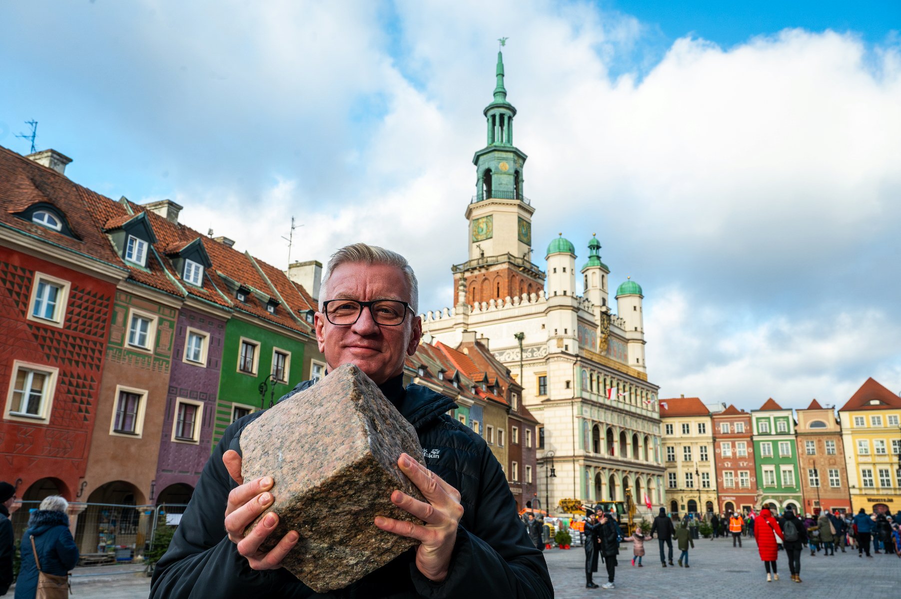 Na pierwszym planie Jacek Jaśkowiak trzymający w dłoniach kamienną kostkę. W tle ratusz i domki budnicze. - grafika artykułu