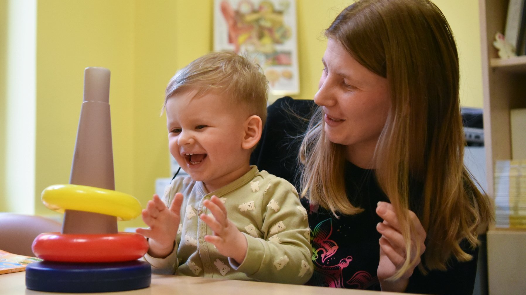 Na zdjęciu uśmiechnięte dziecko na kolanach mamy, patrzące na zabawkę