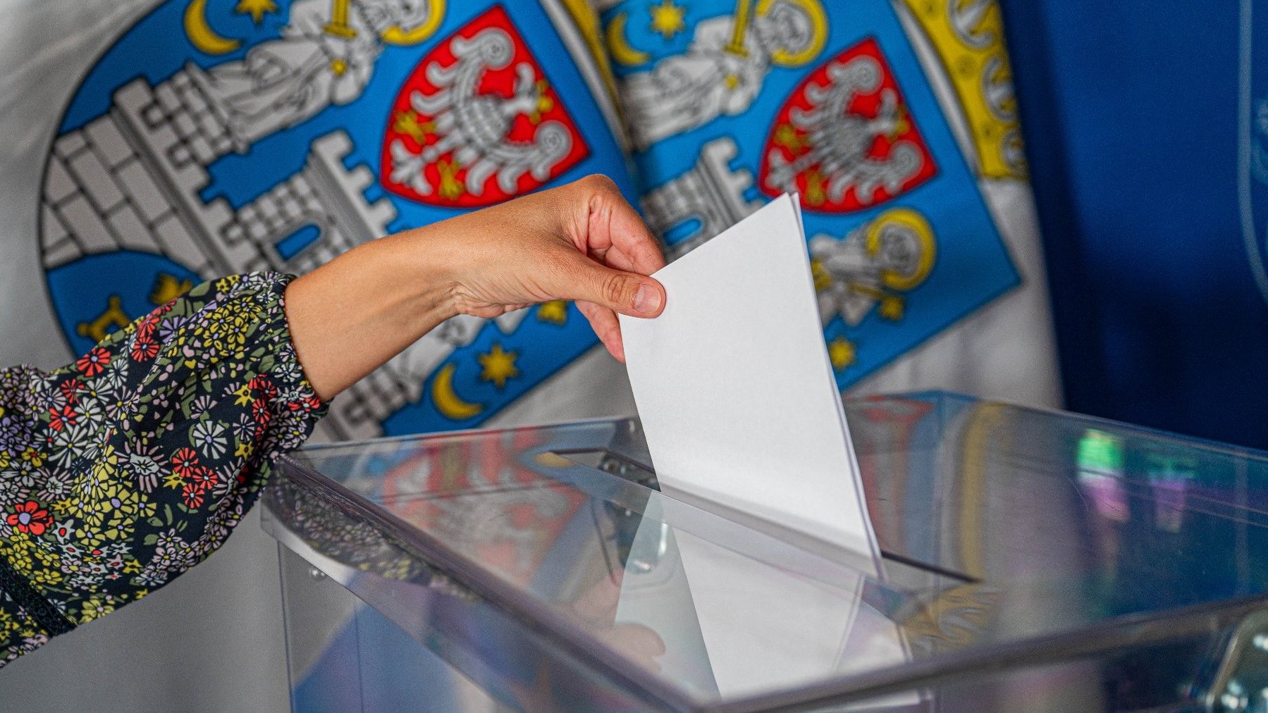 Zdjęcie przedstawia rękę wrzucającą kartę do głosowania do urny wyborczej. W tle flagi z herbem Miasta Poznania.
