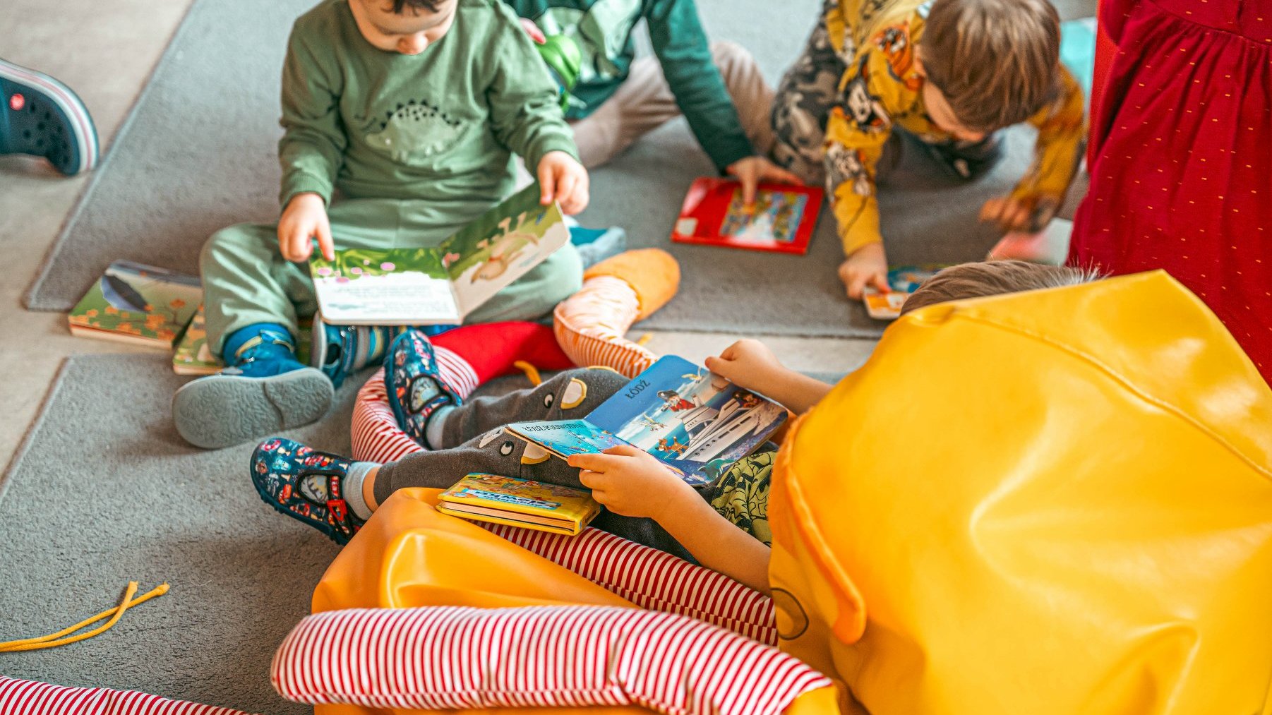 Na zdjęciu dzieci bawiące się na podłodze w żłobku, w rękach trzymają książki, obok leży żółty puf