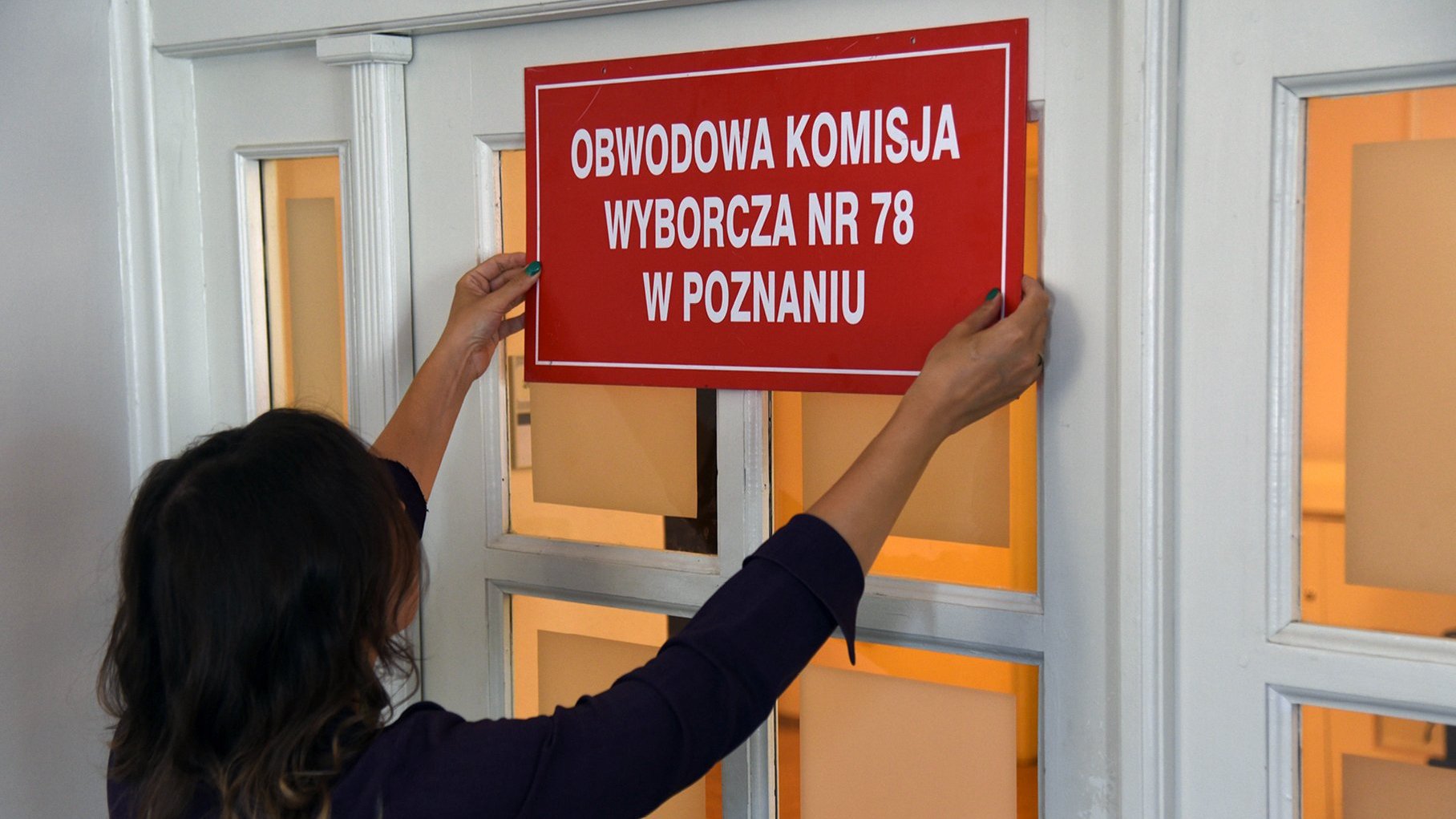 Zdjęcie przedstawia osobę wiesząjącą tabliczkę z napisem "Obwodowa Komisja Wyborcza nr 78 w Poznaniu".
