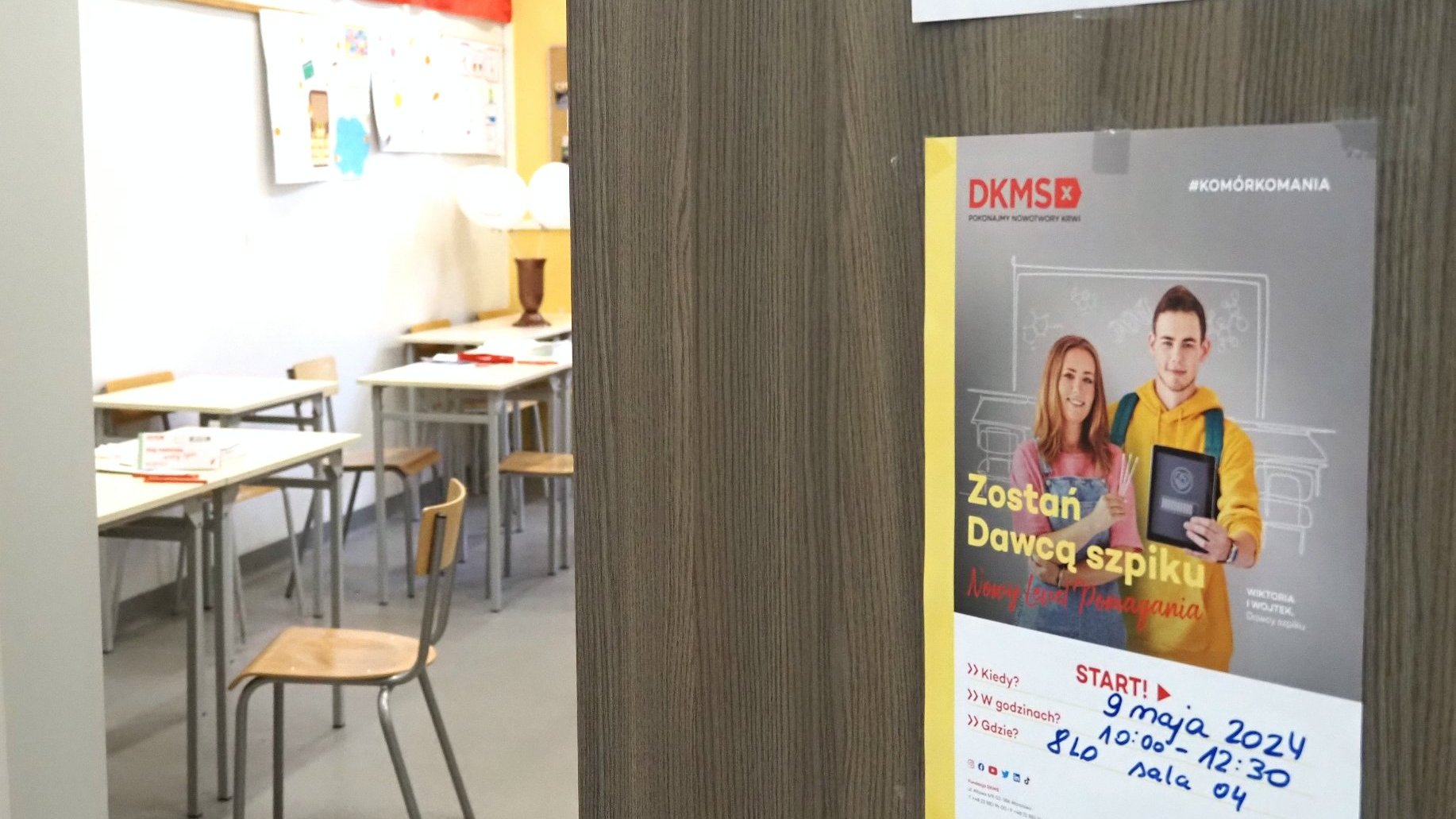 Zdjęcie przedstawia otwarte drzwi do sali lekcyjnej. Na drzwiach wisi plakat z informacją o akcji. Wewnątrz klasy widać ławki i krzesła.