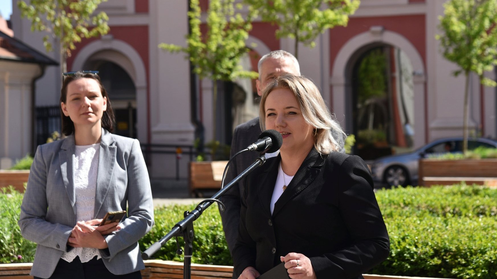 Na zdjęciu trzy osoby na dziedzińcu, dwie kobiety, w tym jedna przy mikrofonie, za nią mężczyzna