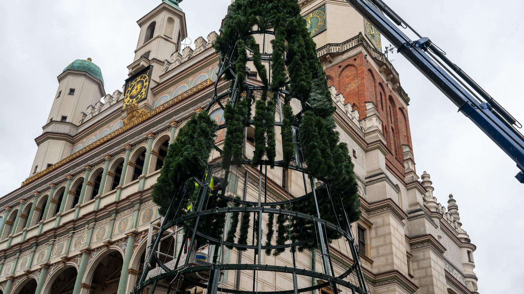 Na środku placu stoi duża, metalowa rama w kształcie choinki, częściowo pokryta zieloną folią. Obok znajduje się dźwig i kilku pracowników, którzy pomagają w montażu choinki. W tle widoczny jest ozdobny budynek z wieżą zegarową i licznymi architektonicznymi detalami, takimi jak łuki i dekoracyjne elementy. Scena wygląda na przygotowania do świątecznego wydarzenia w historycznej lub ważnej lokalizacji