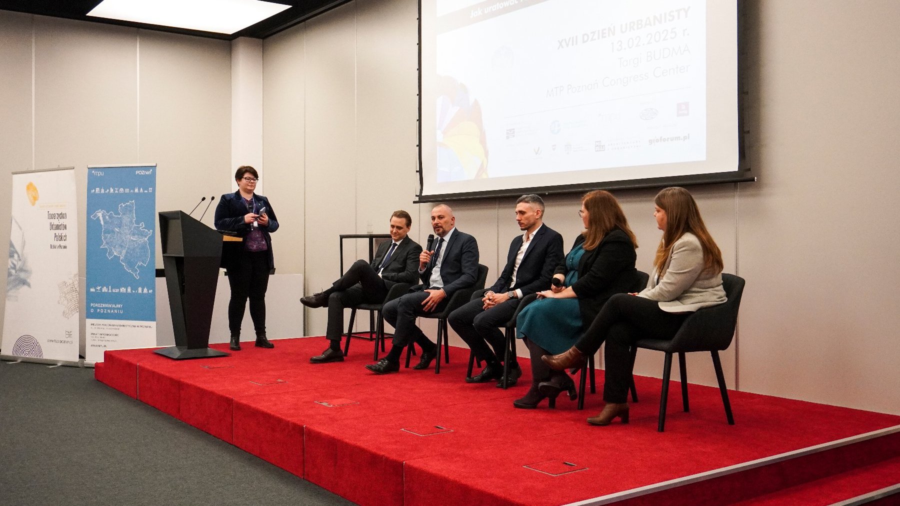 Konferencja urbanistyczna. Na scenie znajduje się sześć osób: jedna osoba stoi przy mównicy po lewej stronie obrazka, pięć osób siedzi na krzesłach na scenie. Osoba przy mównicy mówi do mikrofonu, a siedzące osoby są uczestnikami panelu dyskusyjnego. Za mównicą widoczne są dwa banery: jeden z mapą i tekstem po polsku, drugi z tekstem i grafiką.