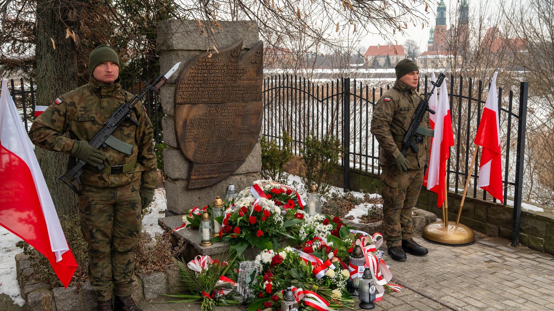 Zdjęcie przedstawia dwóch żołnierzy stojących na straży obok pomnika pamiątkowego. Żołnierze są ubrani w mundury kamuflażowe i trzymają karabiny. Pomnik wykonany jest z kamienia i ma wyryty napis na tabliczce. Wokół pomnika znajduje się wiele kwiatowych wieńców i bukietów, z których wiele jest ozdobionych czerwono-białymi wstążkami, co jest nawiązaniem do kolorów flagi Polski. Obok pomnika ustawiono również kilka flag Polski.