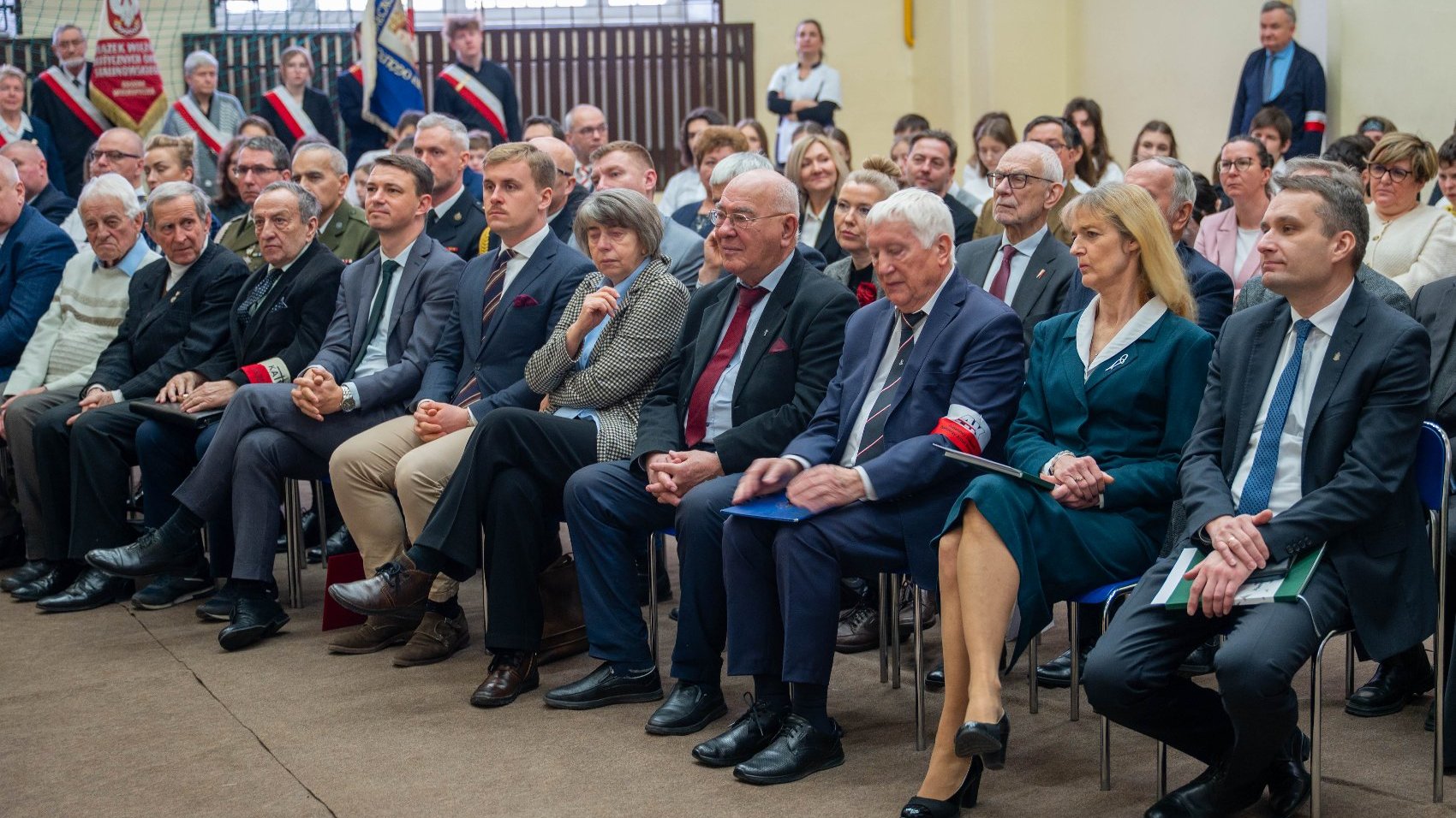 Wnętrze budynku. Grupa ludzi siedzi w rzędach, ubrana w formalne stroje: garnitury i sukienki. Niektórzy z uczestników noszą opaski na rękach. W tle widoczne są flagi oraz banery, co sugeruje, że jest to oficjalne wydarzenie lub ceremonia. Tło zdjęcia obejmuje okna i siatkę, co może wskazywać na halę lub audytorium.