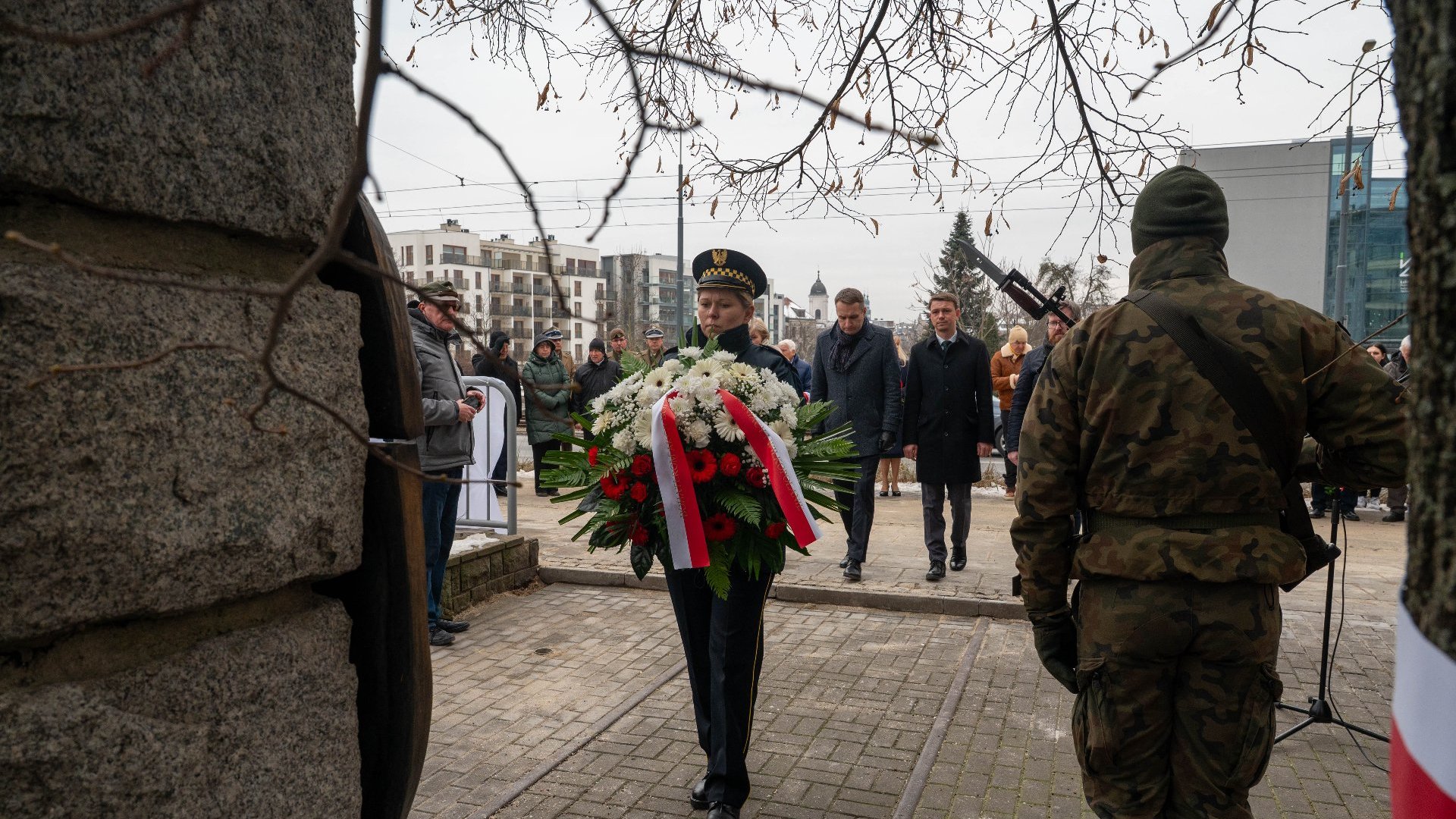 Osoba w mundurze wojskowym lub ceremonialnym niesie duży wieniec kwiatowy ozdobiony czerwonymi i białymi kwiatami oraz wstążkami. Osoba idzie przed grupą ludzi, z których niektórzy są ubrani w formalne stroje.