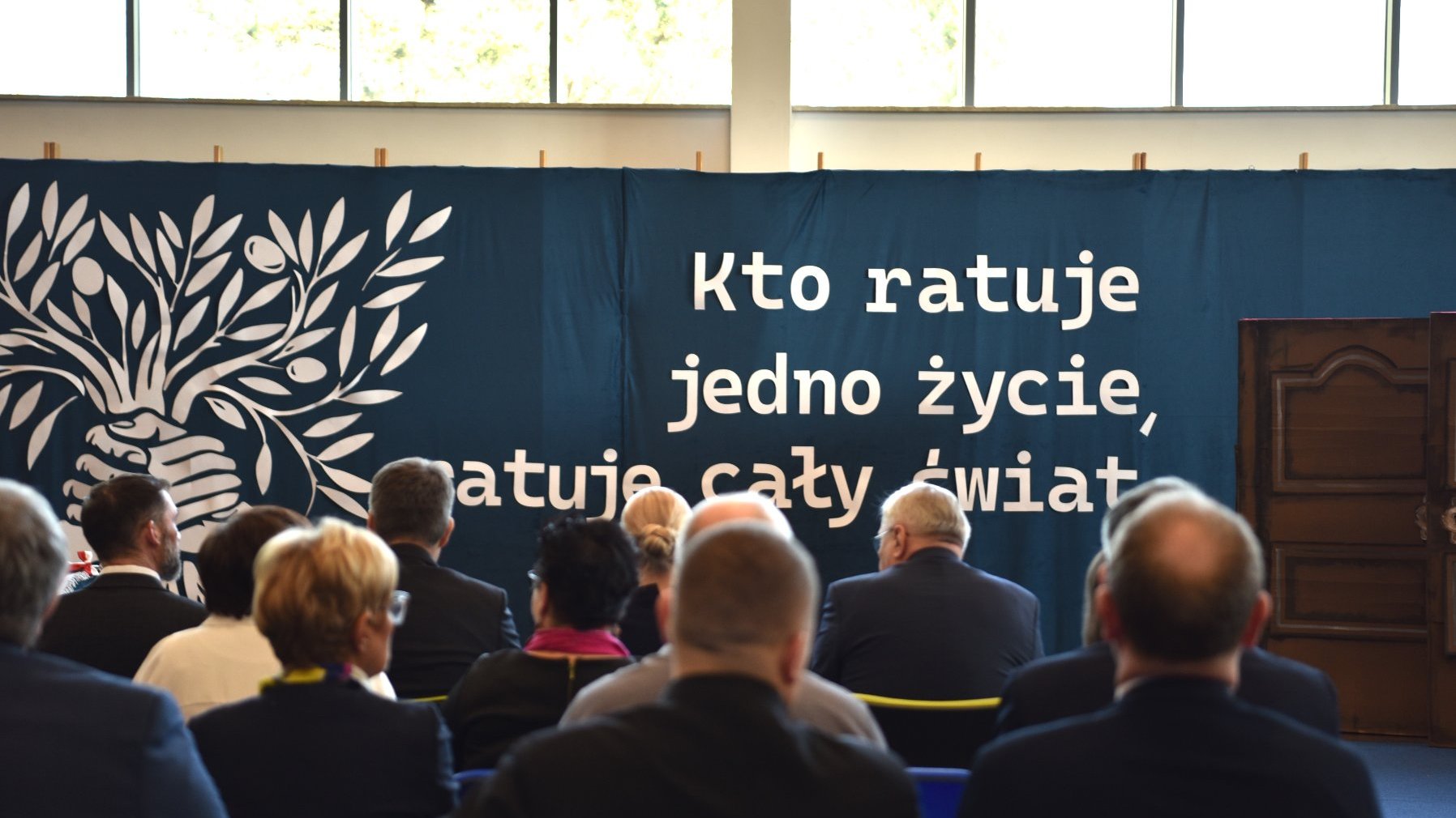 Zdjęcie przedstawia napis "Kto ratuje jedno życie ratuje cały świat" umieszczony na dużej płachcie. Widać też siedzących na widowni ludzi.