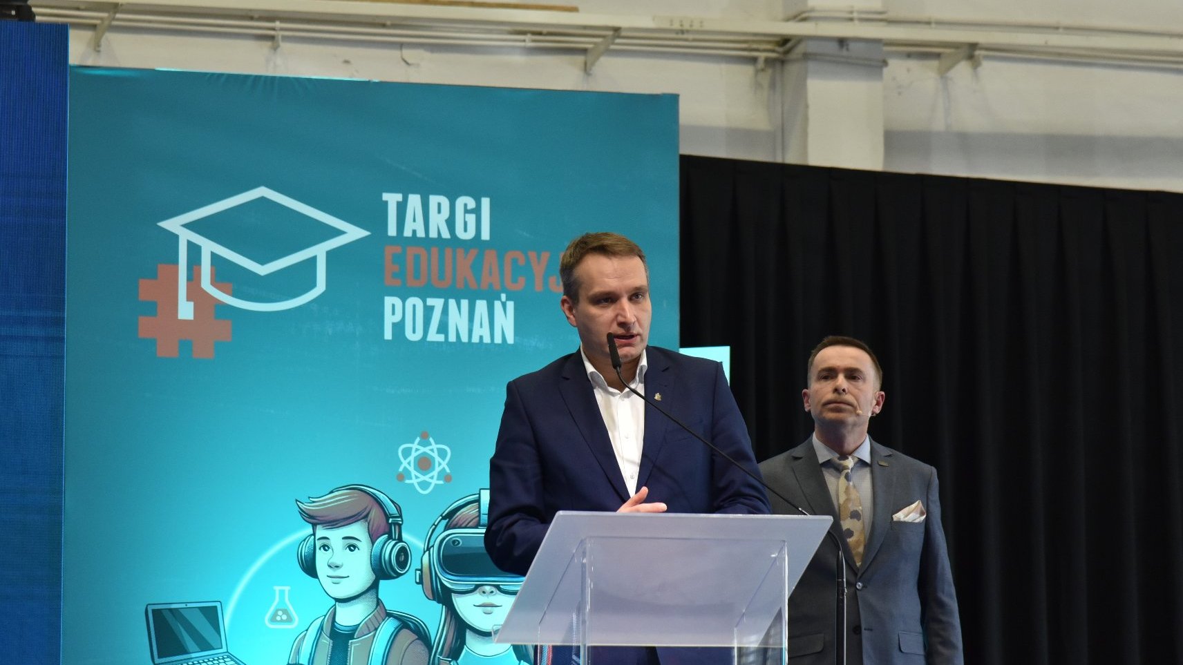 Na zdjeciu dwóch męzczyzn, jeden stoi za mównicą, otwiera Targi Edukacyjne