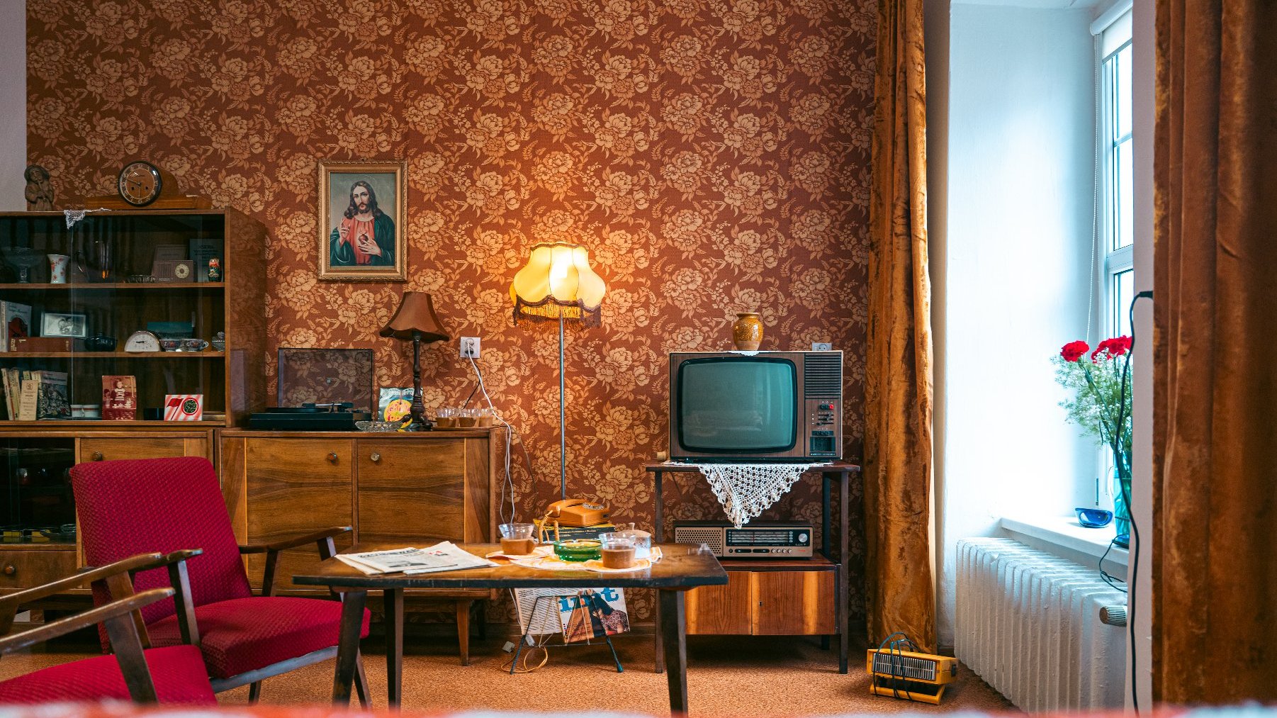 Salon w stylu vintage, z różnymi retro elementami. Pokój ma tapetę w czerwone kwiatowe wzory, drewnianą szafkę z półkami, na których znajdują się różne przedmioty, oraz oprawiony obraz Jezusa na ścianie. Na drewnianym stojaku stoi telewizor w stylu retro, pod którym leży koronkowa serwetka. Obok telewizora znajduje się lampka podłogowa z żółtym abażurem. Przed telewizorem stoi drewniany stolik kawowy z gazetami, szklana popielniczka i kilka innych przedmiotów.