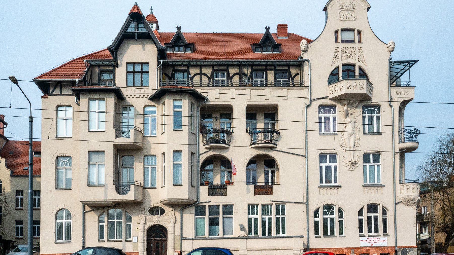 Wielopiętrowy budynek mieszkalny o bogato zdobionej architekturze, łączący różne style, w tym secesję. Na elewacji znajdują się dekoracyjne detale, takie jak misternie rzeźbienia, balkony z kutymi żelaznymi balustradami oraz różnorodne kształty i rozmiary okien. Dach jest stromo spadzisty z lukarnami i szczytami. Przed budynkiem zaparkowanych jest kilka samochodów, widoczne są latarnia uliczna i linie energetyczne.