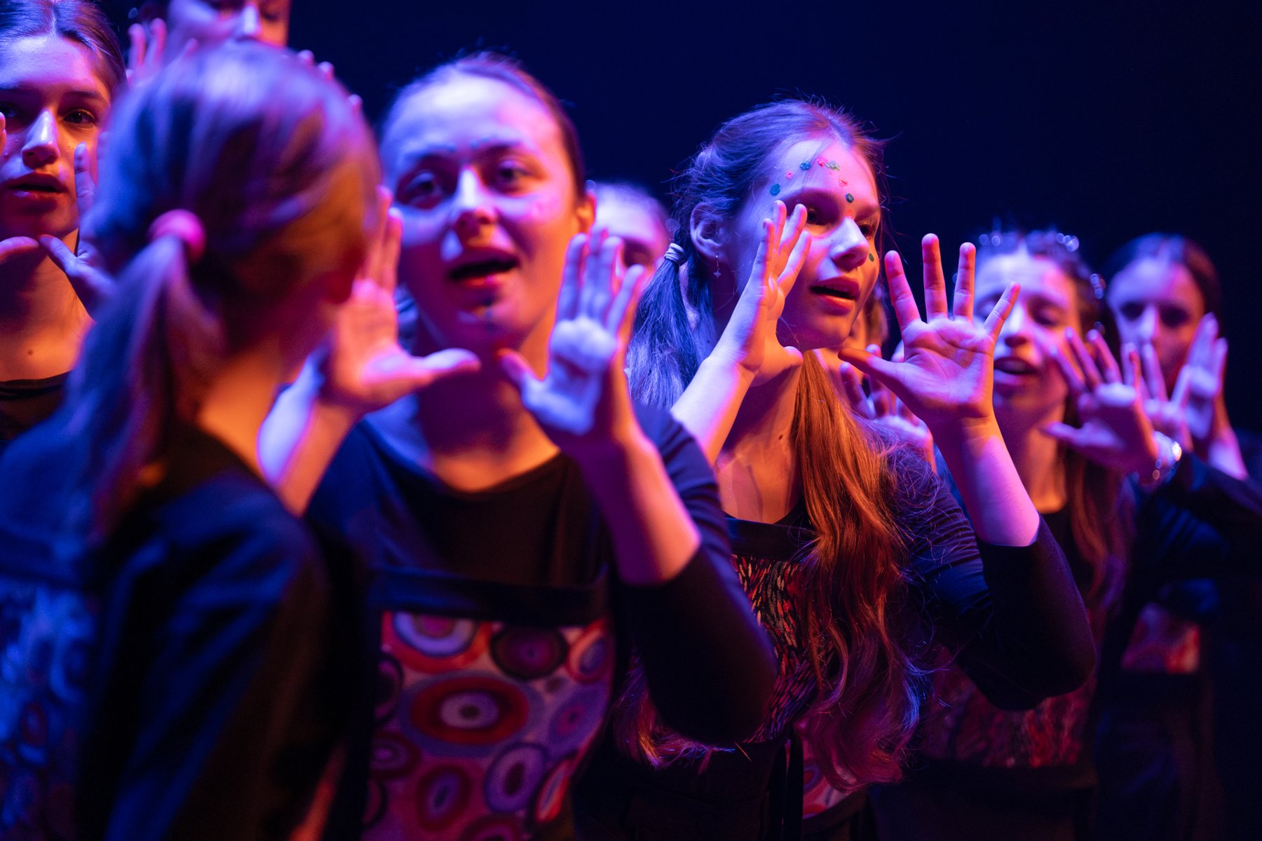 Scena oświetlona w tonacji niebiesko-fioletowej. Grupa osób stoi blisko siebie, wykonując gesty rękami. Część osób ma na sobie wzorzyste ubrania. - grafika artykułu