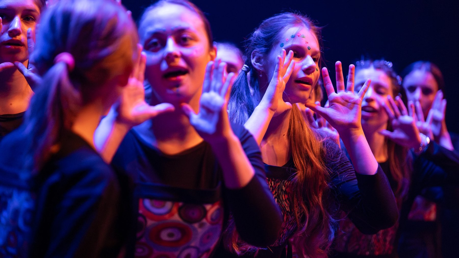 Scena oświetlona w tonacji niebiesko-fioletowej. Grupa osób stoi blisko siebie, wykonując gesty rękami. Część osób ma na sobie wzorzyste ubrania.