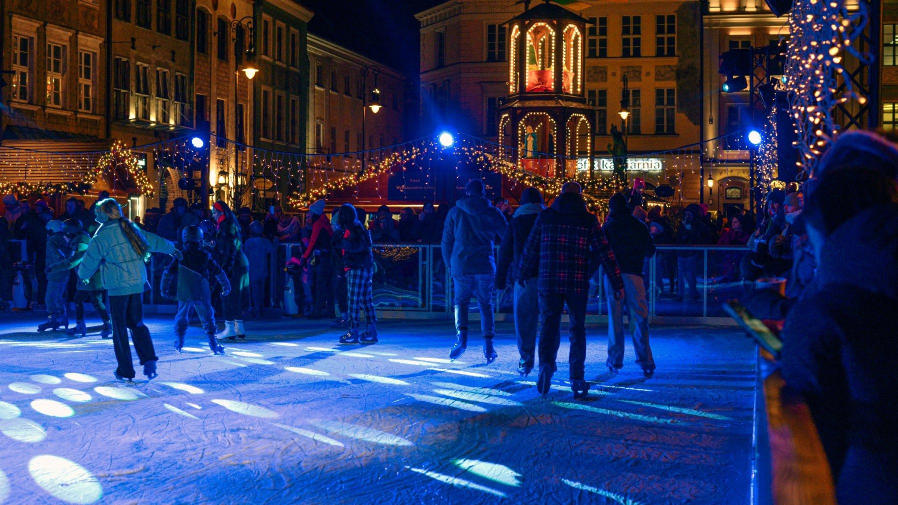 Zdjęcie przedstawia ludzi jeżdżących na łyżwach po lodowisku. W tle widać budynki znajdujące się na rynku.