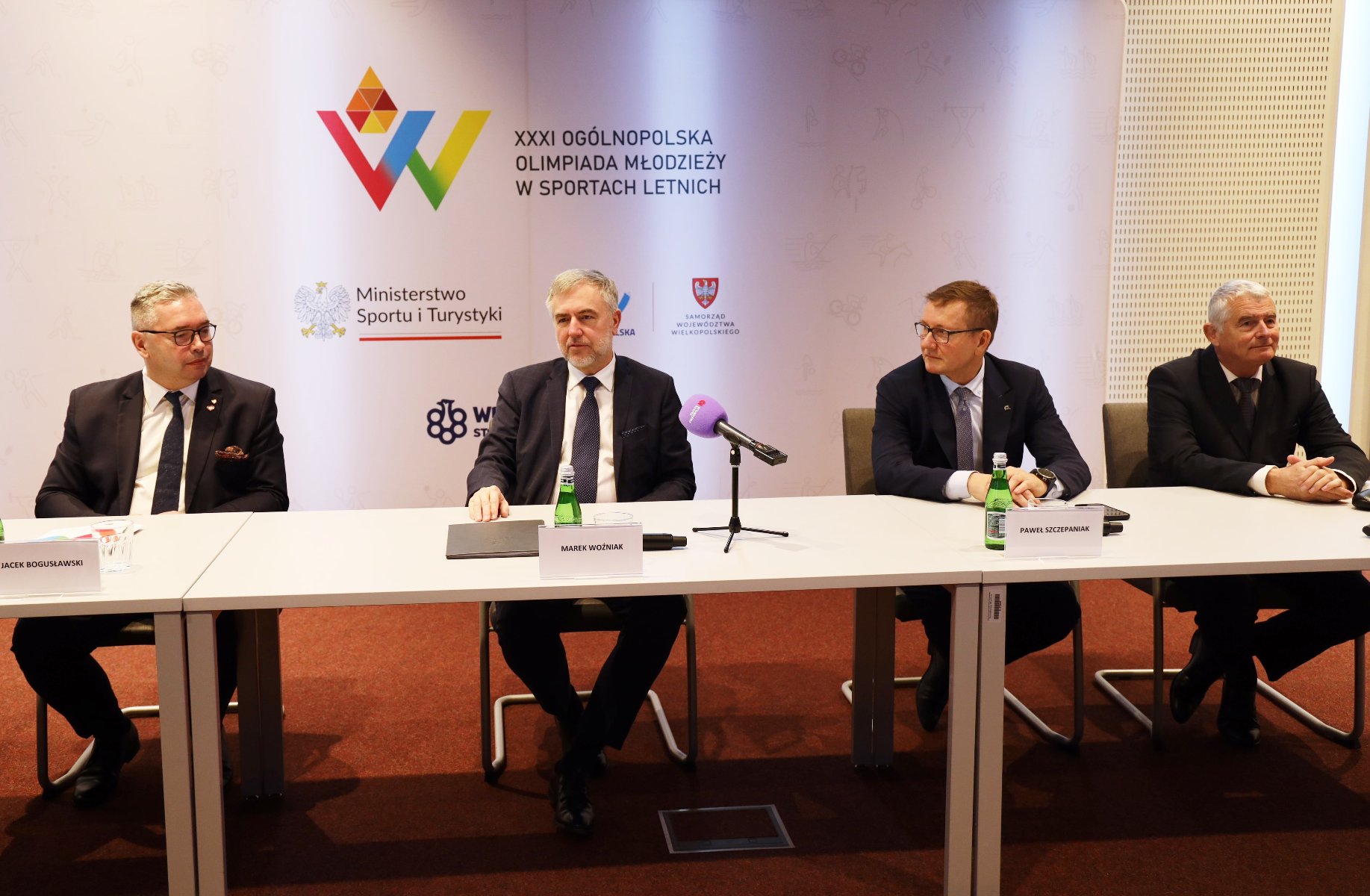 Konferencja prasowa przed Ogólnopolską Olimpiadą Młodzieży w sportach letnich Wielkopolska 2025 - za stołem konferencyjnym siedzi 4 mężczyzn w garniturach. Od lewej są to: Jacek Bogusławski - członek zarządu Województwa Wielkopolskiego, Marek Woźniak - Marszałek Województwa Wielkopolskiego, Paweł Szczepaniak - prezes Wielkopolskiego Stowrzyszenia Sportowego, Tomasz Wiktor - dyrektor Departamentu Sportu i Turystyki Urzędu Marszałkowskiego Województwa Wielkopolskiego - grafika artykułu