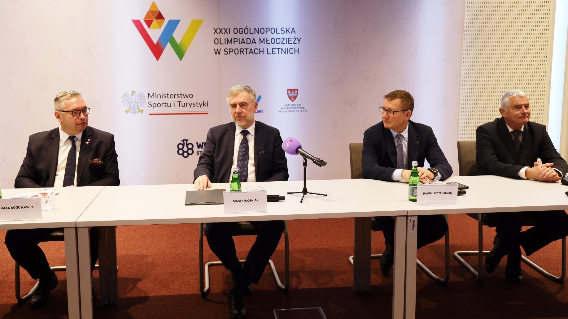 Konferencja prasowa przed Ogólnopolską Olimpiadą Młodzieży w sportach letnich Wielkopolska 2025 - za stołem konferencyjnym siedzi 4 mężczyzn w garniturach. Od lewej są to: Jacek Bogusławski - członek zarządu Województwa Wielkopolskiego, Marek Woźniak - Marszałek Województwa Wielkopolskiego, Paweł Szczepaniak - prezes Wielkopolskiego Stowrzyszenia Sportowego, Tomasz Wiktor - dyrektor Departamentu Sportu i Turystyki Urzędu Marszałkowskiego Województwa Wielkopolskiego