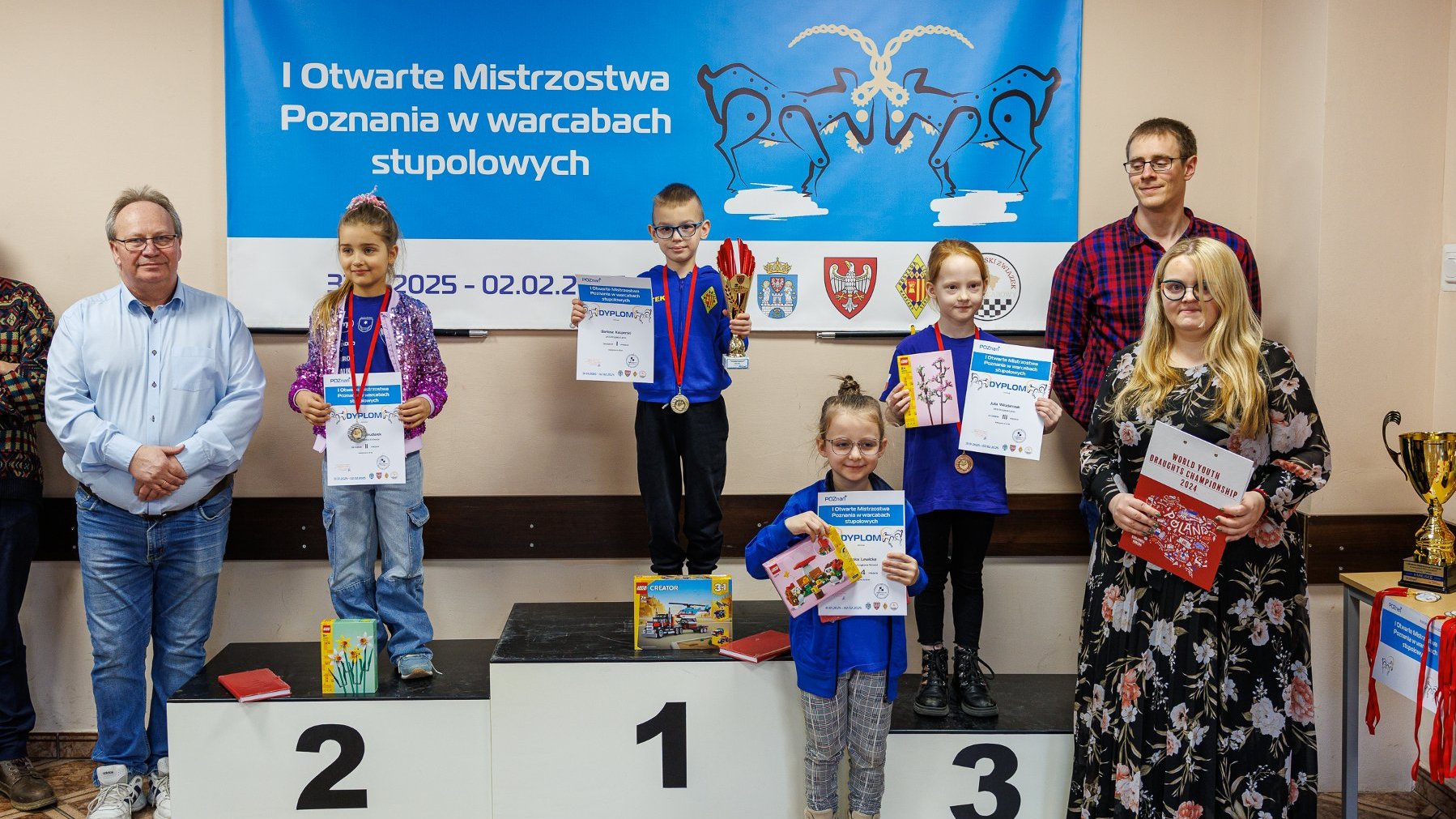 I Otwarte Mistrzostwa Poznania w warcabach stupolowych - na podium stoją najlepsi zawodnicy w kategorii do 8 lat, w tym jeden chłopiec i trzy dziewczynki, trzymając dyplomy, medale i nagrody. Oprócz nich stoi prowadząca wydarzenie oraz organizatorzy, W tle baner imprezy z nazwą, datą, logotypem organizatorów oraz koziołków