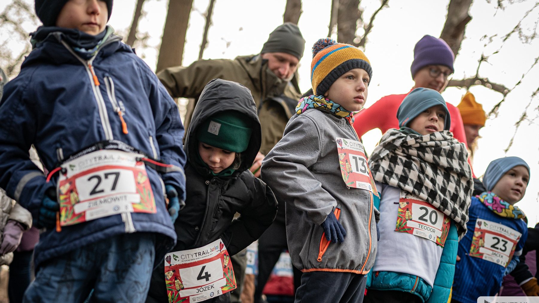 Bieg City Trail w Poznaniu, fot. Piotr Oleszak - dzieci w zimowych strojach, w czapkach i rękawiczach, z zawieszonymi numerami startowymi rozgrzewają się przed biegiem w lesie