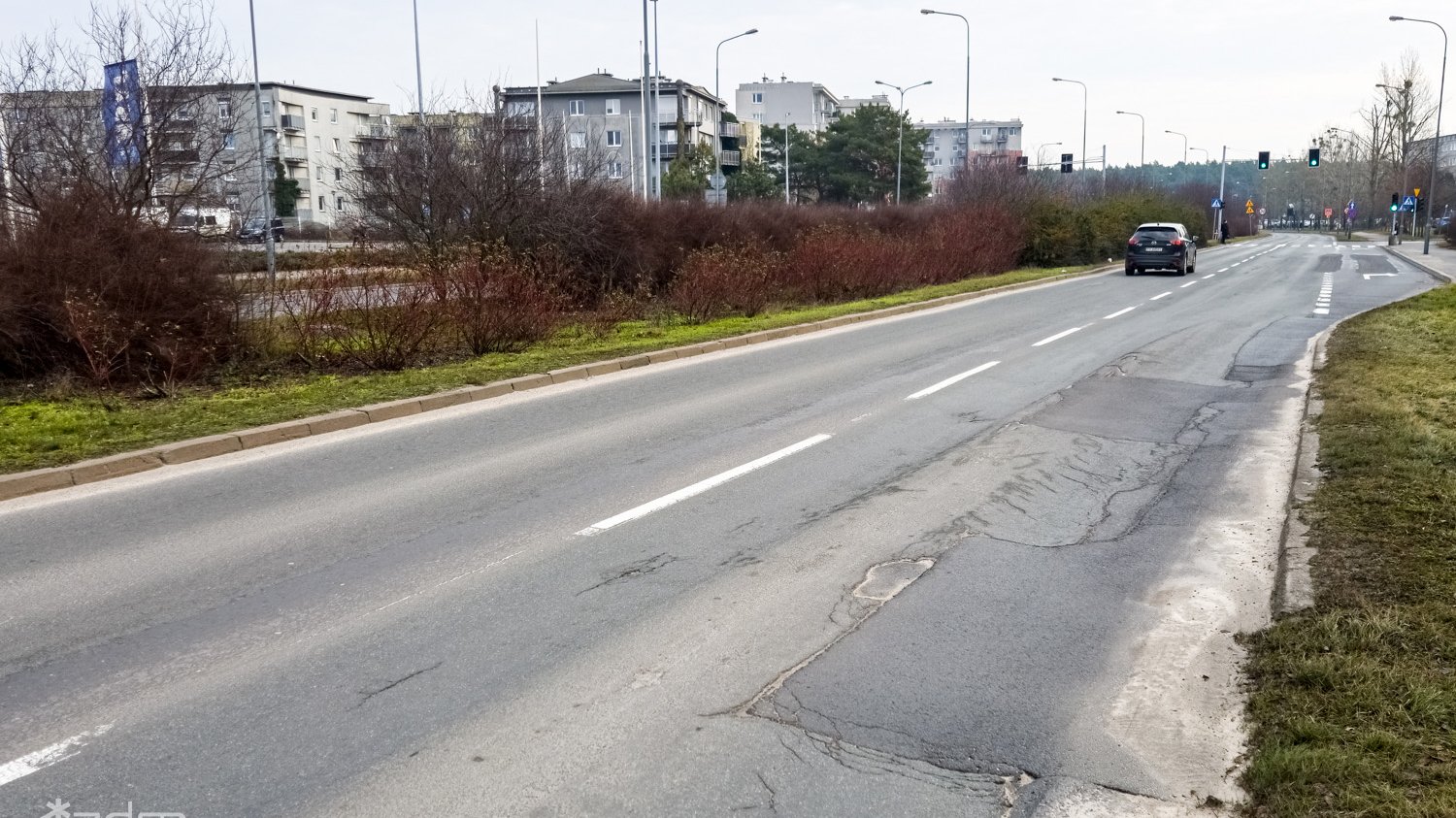 Zniszczona nawierzchnia ul. Opieńskiego i pas zieleni między jezdniami. W tele za ulicą bloki mieszkalne.