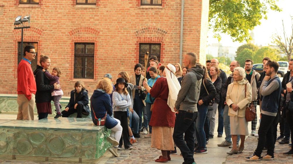 Grupa zwiedzających słucha opowieści przewodnika. Za plecami ludzi budynek z czerwonej cegły, po lewej szklana instalacja.