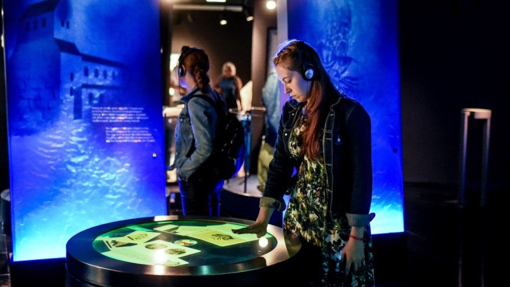 Dwie kobiety w słuchawkach od audiobooka zwiedzają ekspozycję. Jedna z nich dotyka okrągłego ekranu ręką. Instalacja świeci niebieskim i zielonkawym światłem.