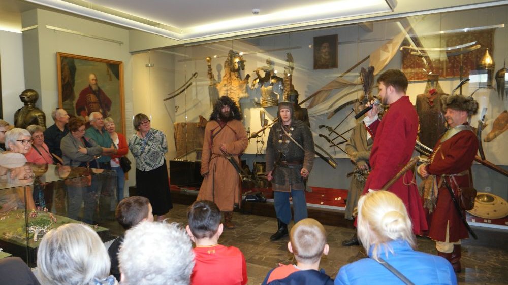 Mężczyźni przebrani w stroje średniowiecznych wojów i stroje szlachciców oprowadzają grupę zwiedzających po wystawie broni dawnej.