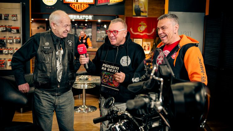 Trójka mężczyzn stoi w studiu z logami Hard Rock Cafe, Harley Davidson i Antyradio. Śmieją się i rozmawiają, Jurek Owsiak w bluzie orkiestry stoi w środku.