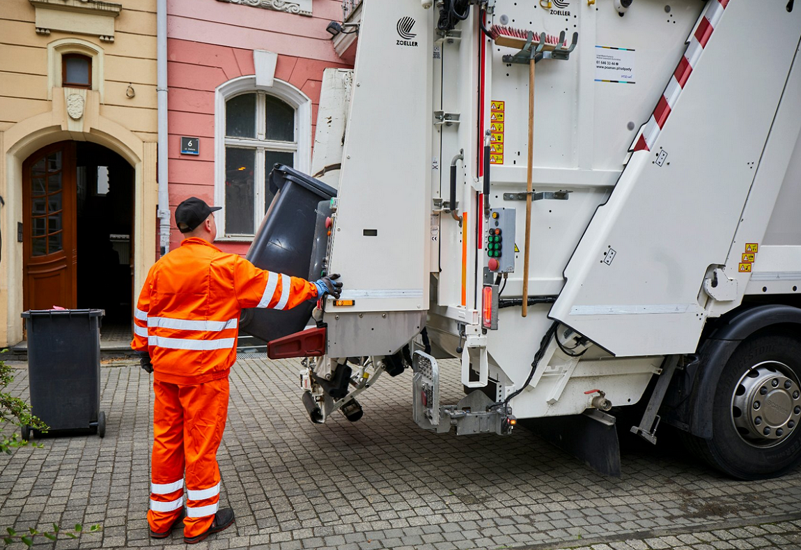 samochód śmieciarka stoi przed kamienicą, obok samochodu pracownik wirmy wywożącej odpady w roboczym kombinezonie - grafika artykułu