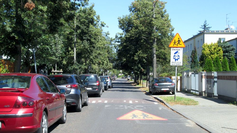 Zdjęcie przedstawia ulicę, na obrzeżach której stoją zaparkowane samochody. Widać na nim także znak "Uwaga, przejście dla dzieci"