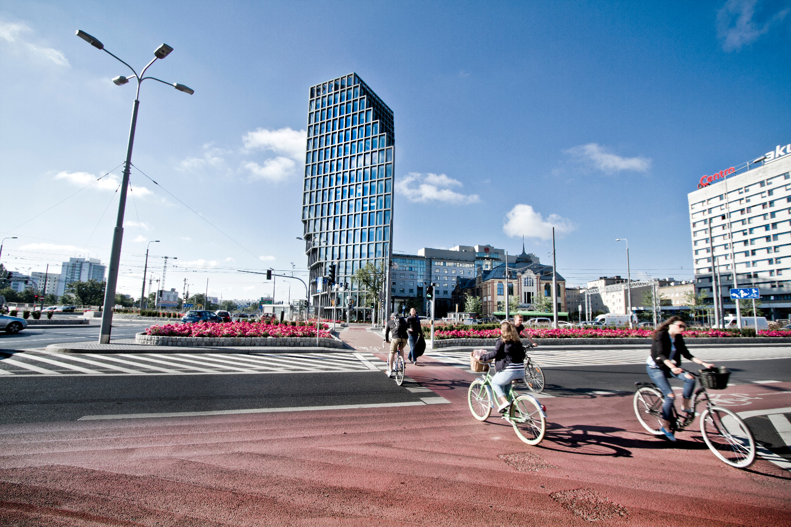 Osoby jadące na rowerach przez rondo, na drugim planie różowe kwiaty i nowoczesne budynki. - grafika artykułu