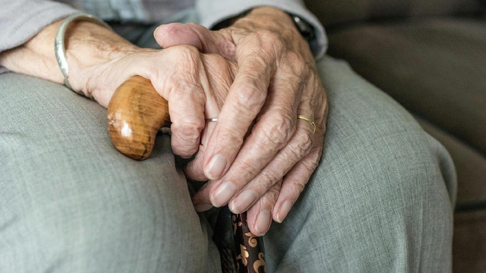Zdjęcie: dłonie starszego człowieka, wsparte na kolanach, trzymające laskę