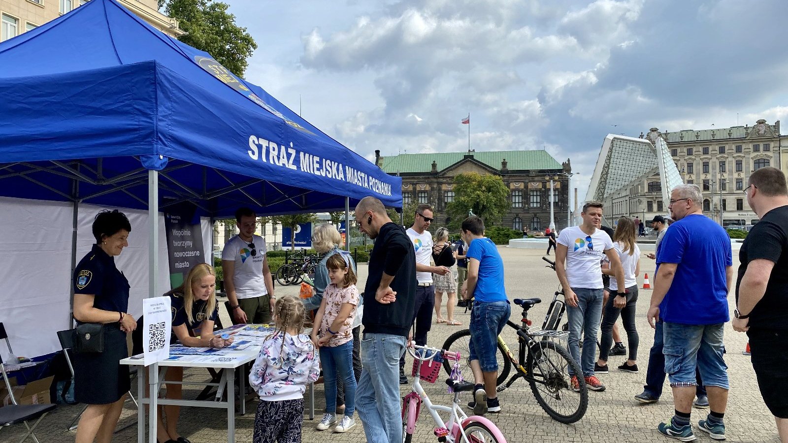 Konsultacje z mieszkańcami, po prawej stronie niebieski namiot, wszyscy stoją na placu, dwie osoby trzymają rowery, w tle fontanna.