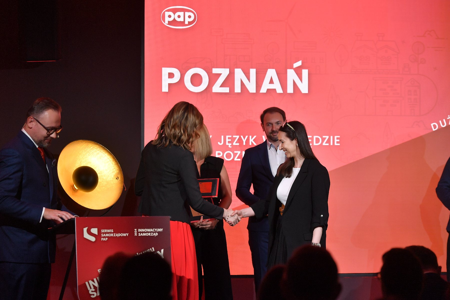 Na pierwszym planie dwie kobiety uściskują dłonie. Jedna z nich wręcza drugiej nagrodę. Za nimi stoją 3 osoby. W tle duży czerwony plakat, na którym wyświetla się napis "Poznań". - grafika artykułu