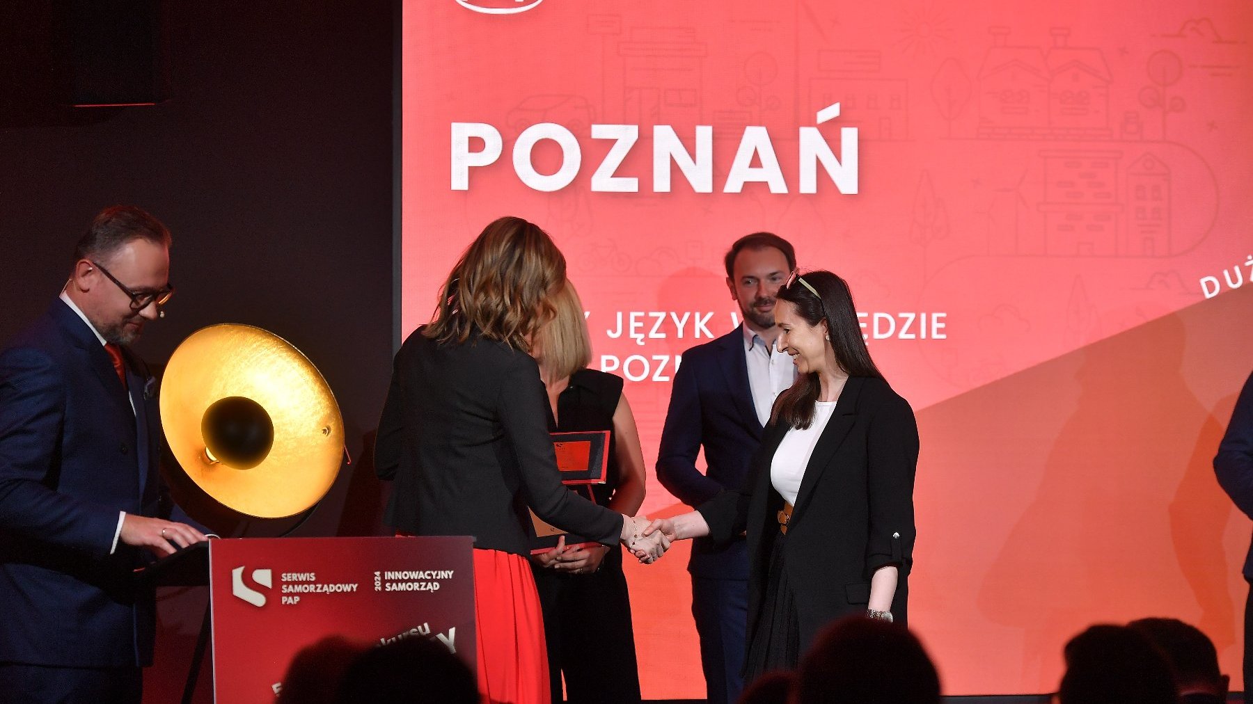 Na pierwszym planie dwie kobiety uściskują dłonie. Jedna z nich wręcza drugiej nagrodę. Za nimi stoją 3 osoby. W tle duży czerwony plakat, na którym wyświetla się napis "Poznań".