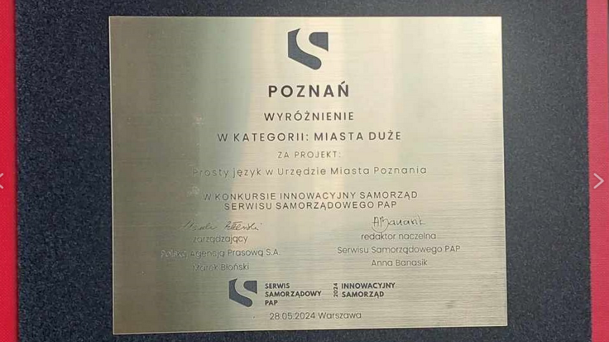 Złota tabliczka z wyróżnieniem w kategorii "Miasta Duże" za projekt. Na tabliczce znajdują się podpisy i loga organizatorów. Tabliczka leży na czarnej gąbce i w czerwonym opakowaniu.