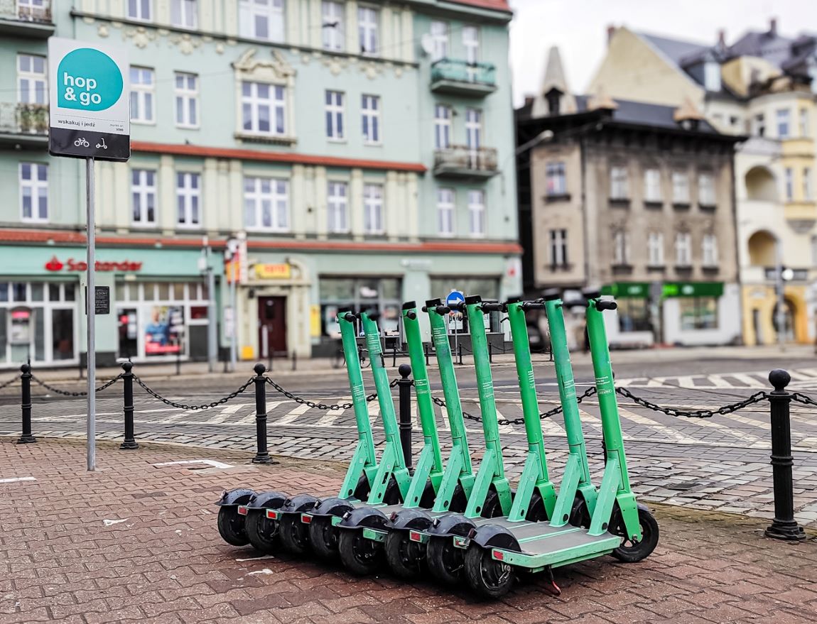 hulajnogi ustawione w rzędzie na chodniku, obok tabliczki z napisem hop&go, za nimi barierki z łańcuchem, ulica, za nią kamienice - grafika artykułu