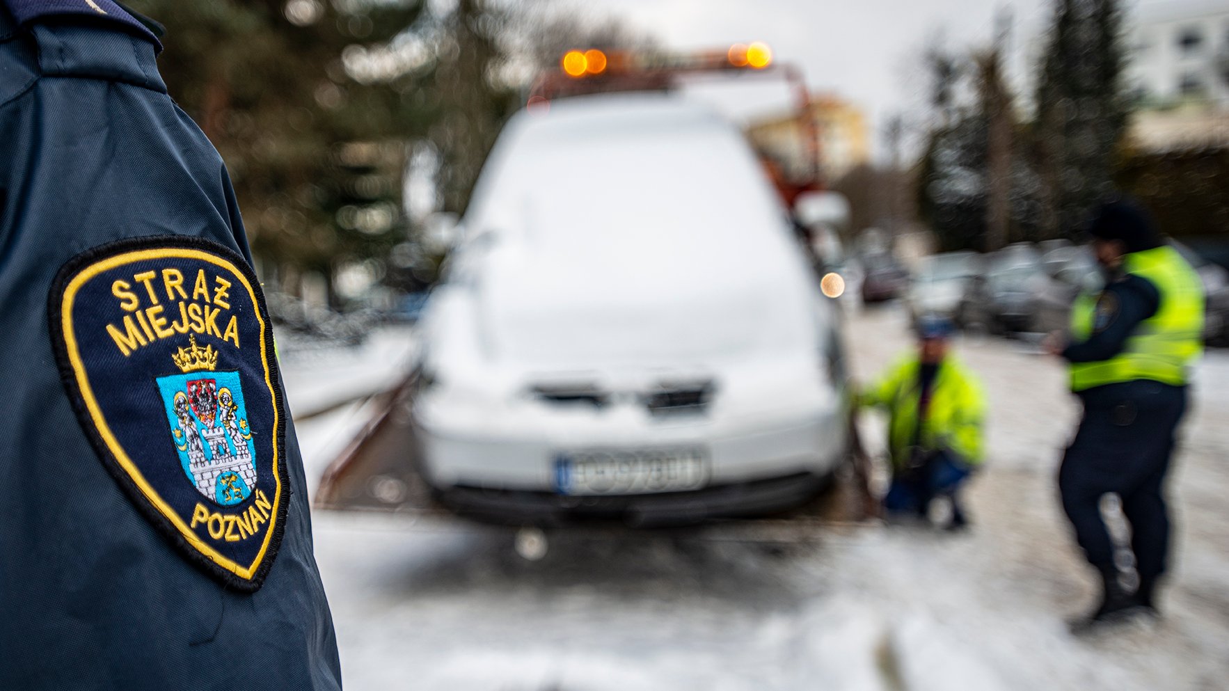 Zdjęcie przedstawia rękaw kurtki strażnika miejskiego. Widać na nim naszywkę z herbem Poznania oraz napisem Straż Miejska Poznań. W tle widać strażników miejskich oraz samochód.