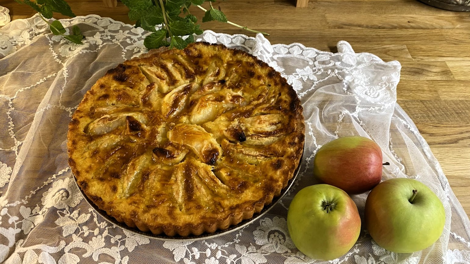 Tarta z jabłkami i trzy jabłka w całości leżą na drewnianym blacie i koronkowym obrusie. - grafika artykułu