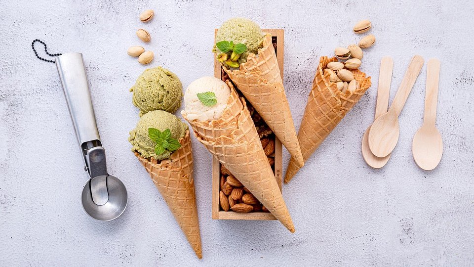 Lody pistacjowe w wafelkach, obok rozsypane pistacje w łupinkach, srebrna łyżka do lodów i drewniane łyżeczki.