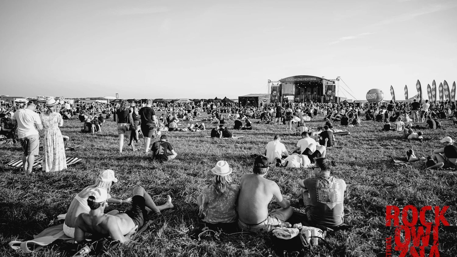 Czarno-białe zdjęcie. Dzień. W oddali scena Rockowizmy. Wokół sceny wiele ludzi siedzi na trawie. Inni stoją i słuchają muzyki.