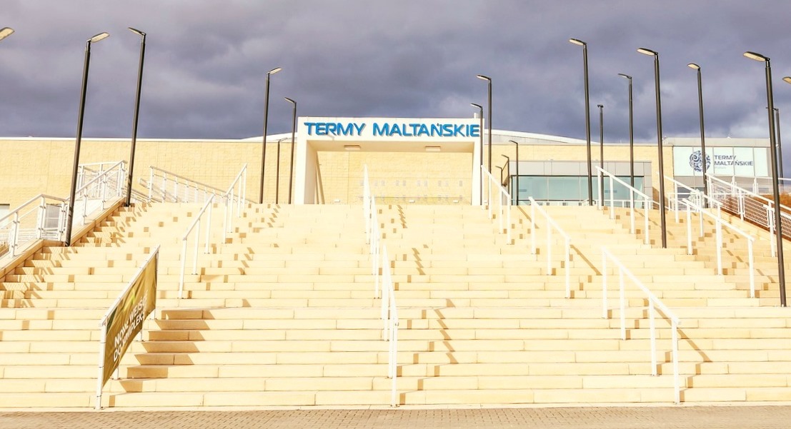 Fot. Termy Maltańskie wejście do budynku Term Maltańskich, wysokie, szerokie schody, na ich szczycie wejście z dużym napisem nad nim TERMY MALTAŃSKIE, na schoda stoją latarnie