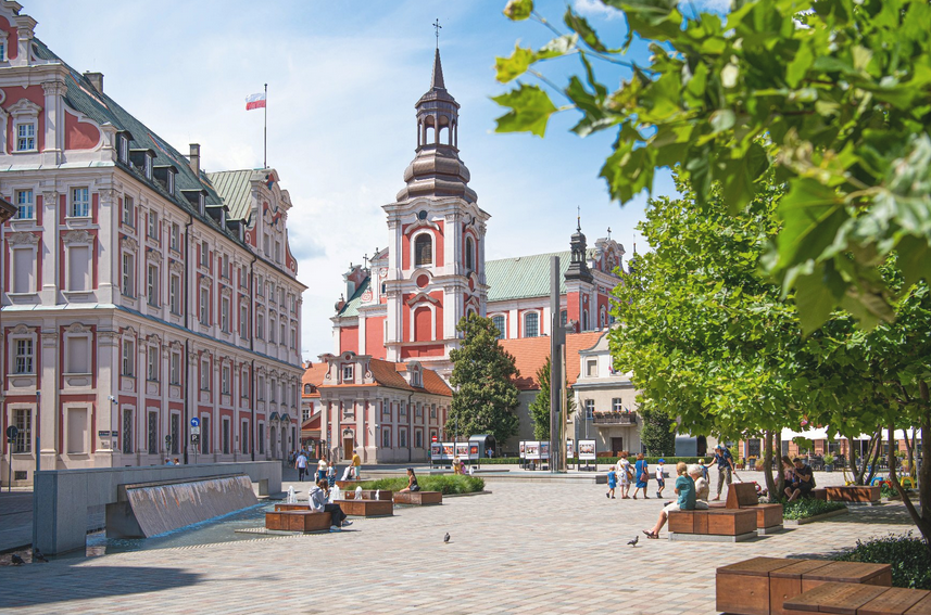 Fot. Miasto Poznań wybrukowany skwer, ludzie siedzą na ławkach lub spacerują, w tle wzniosłe gmachy, na jednym z nich powiewa biało-czerwona flaga
