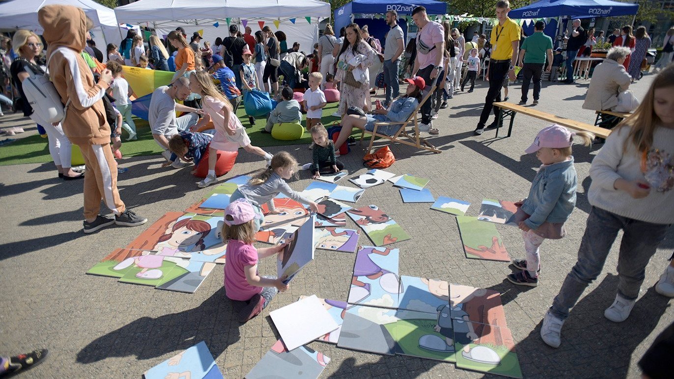 Na zdjęciu grupa ludzi - dzieci i dorośli - układają duże puzzle na ziemi