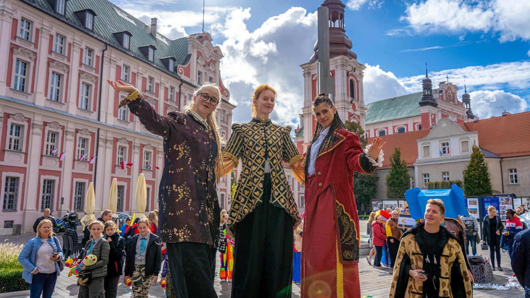 Troje szczudlarzy w kostiumach pozuje do zdjęcia na tle budynku urzędu miasta przy pl. Kolegiackim.