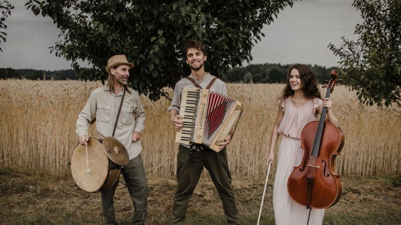 Na zdjęciu trójka muzyków z instrumentami (dwaj mężczyźni oraz kobieta) na tle pola ze zbożem
