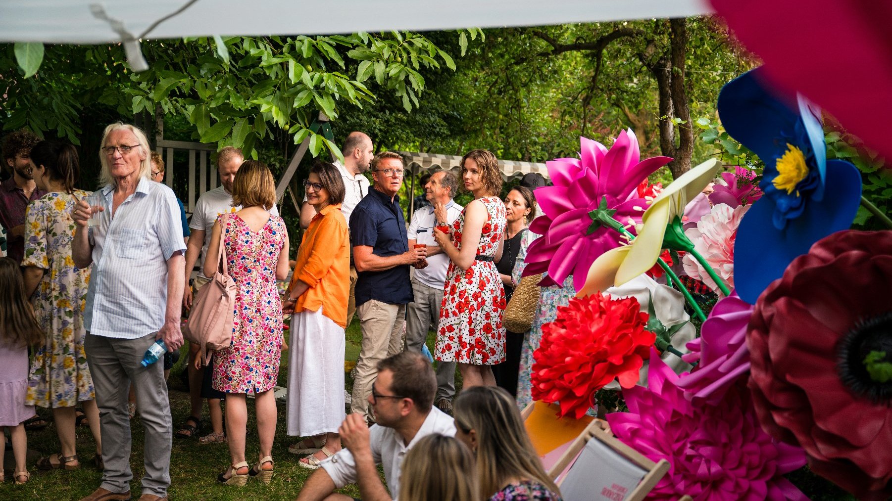 Na zdjęciu grupa ludzi w ogrodzie, obok kolorowe kwiaty