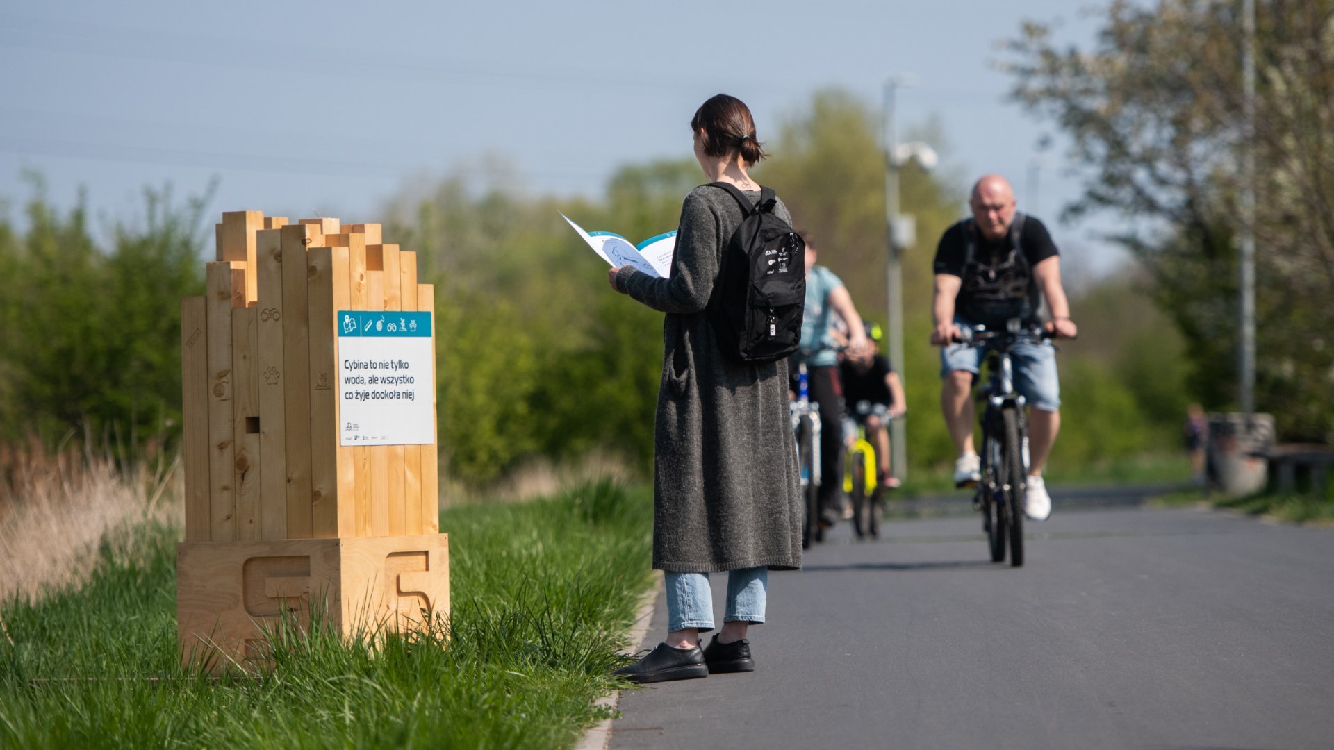 Zdjęci przedstawia totem na ścieżce edukacyjnej. Obok niego stoi osoba, która czyta broszurę poświęconą ścieżce. W tle widać rowerzystów.