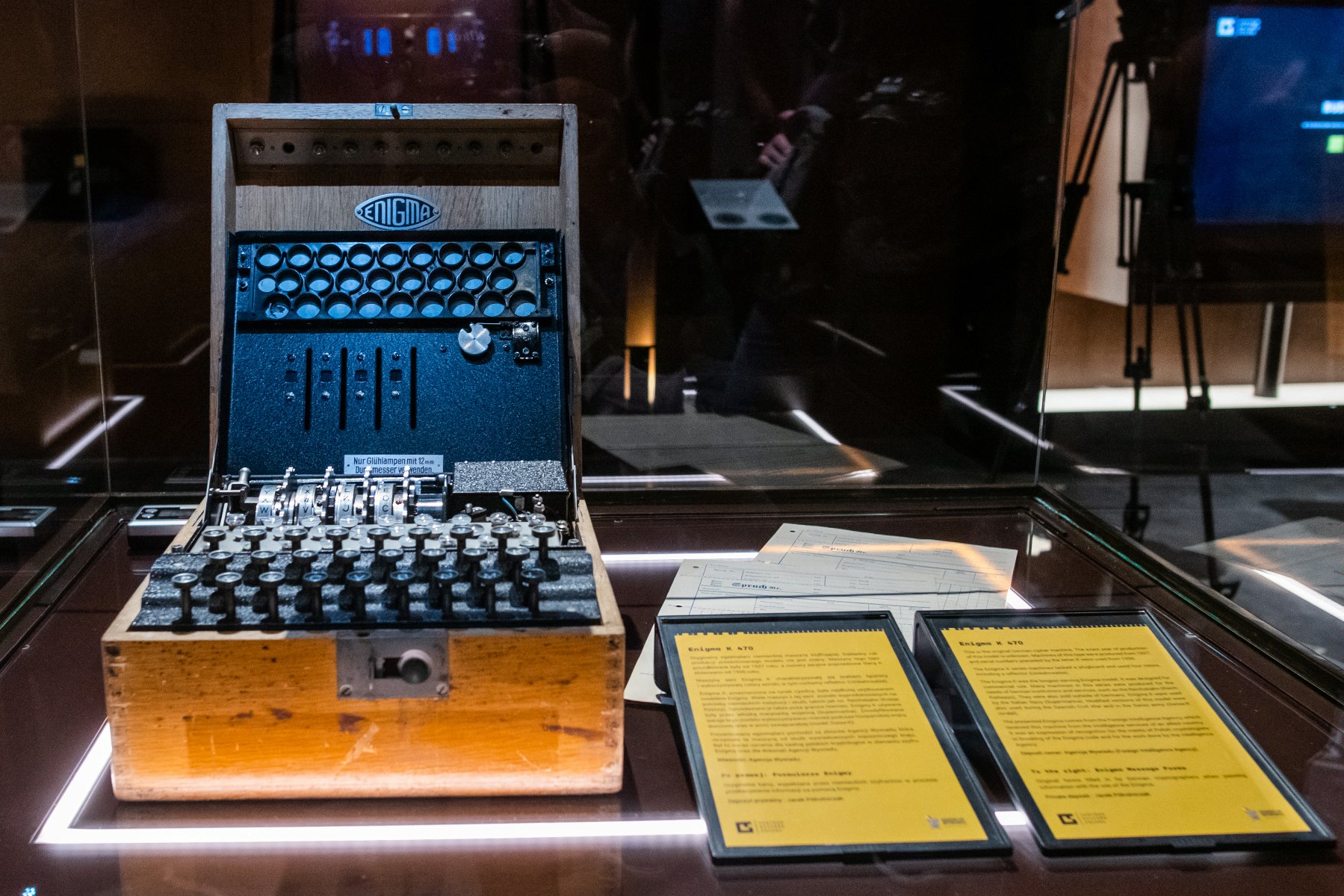 Zdjęcie przedstawia niemiecką maszynę szyfrującą Enigma umieszczoną w gablocie. Obok urządzenia widać dwie tabliczki informacyjne z opisem. - grafika artykułu